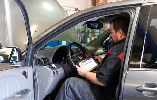 Mechanic using a tablet diagnostic tool inside a car with the hood open in a repair shop.