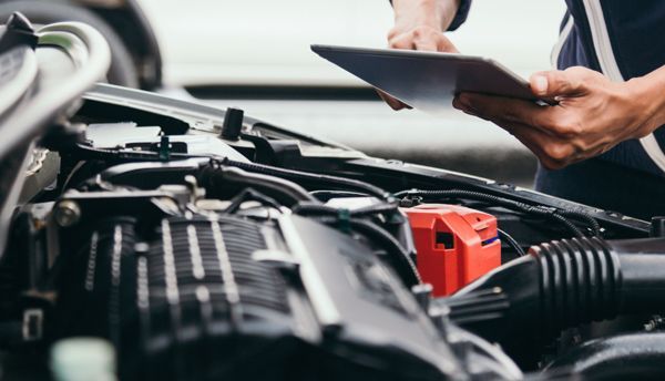Person using a tablet to inspect a car engine; red battery, black engine parts, outdoor setting.