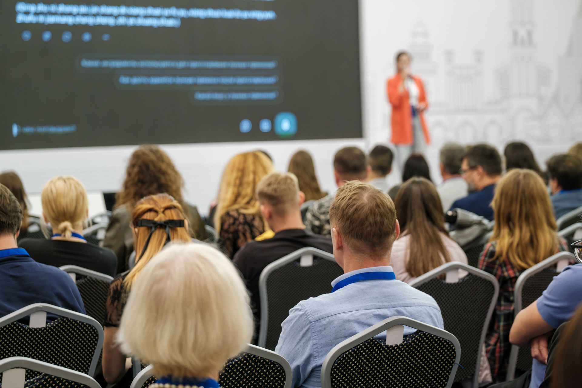 Audience seated facing a presenter speaking on stage beside a large screen during a conference