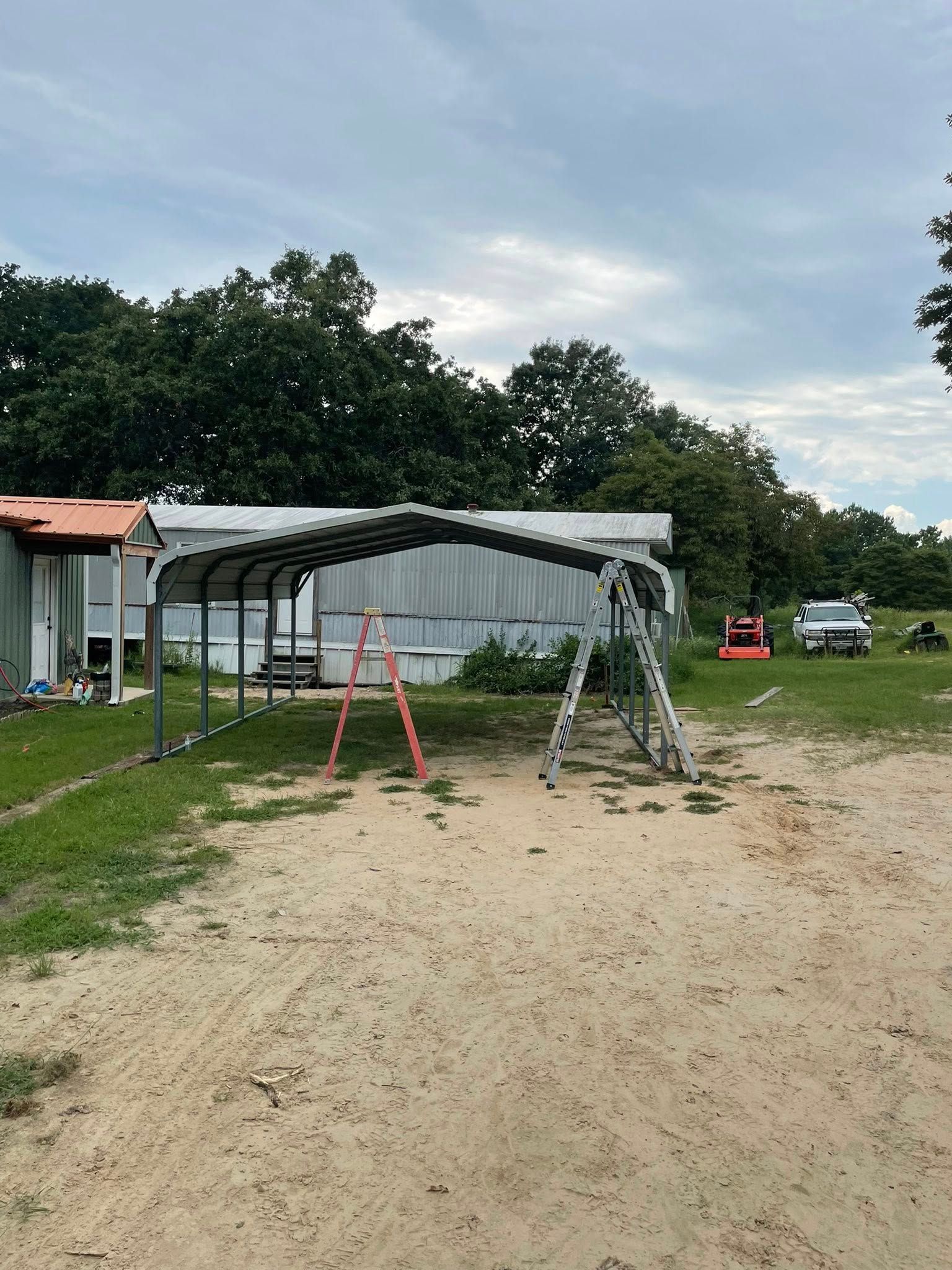 A metal carport under construction next to a mobile home, with ladders and tools visible.