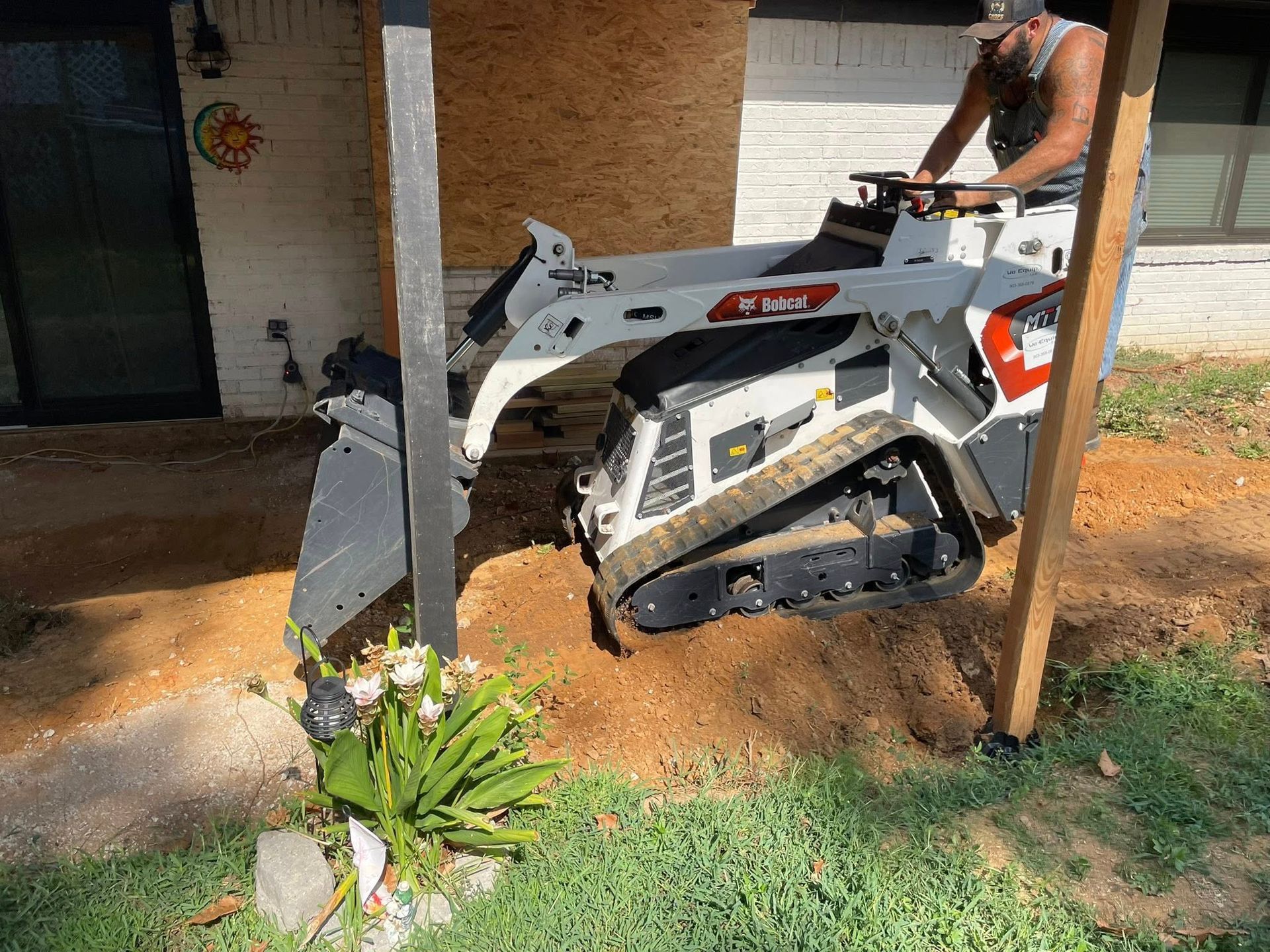 Bobcat skid-steer next to a wooden post, digging in dirt, with a person operating it outdoors.