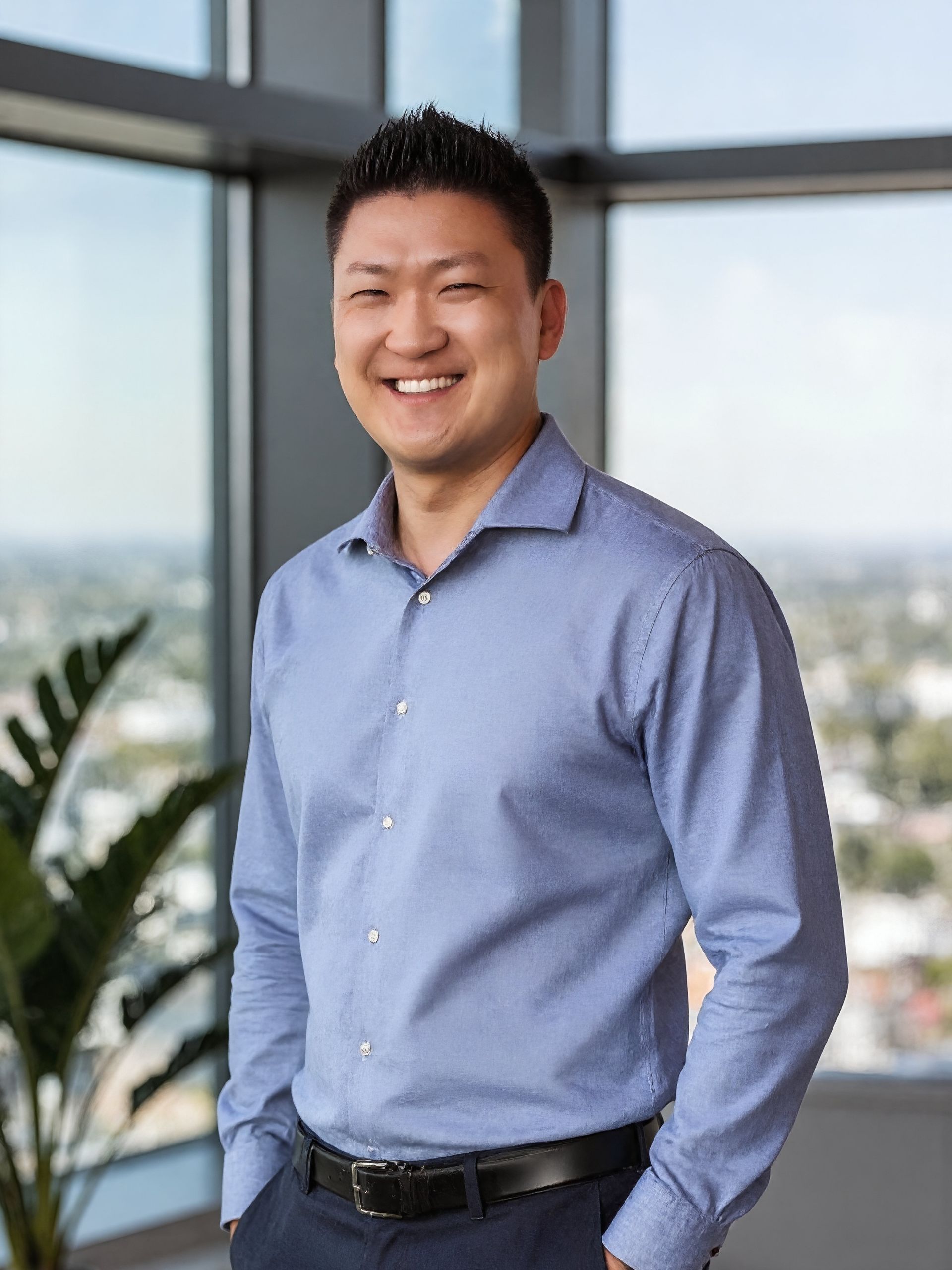 Smiling Asian man in a black suit, black shirt against a white background.