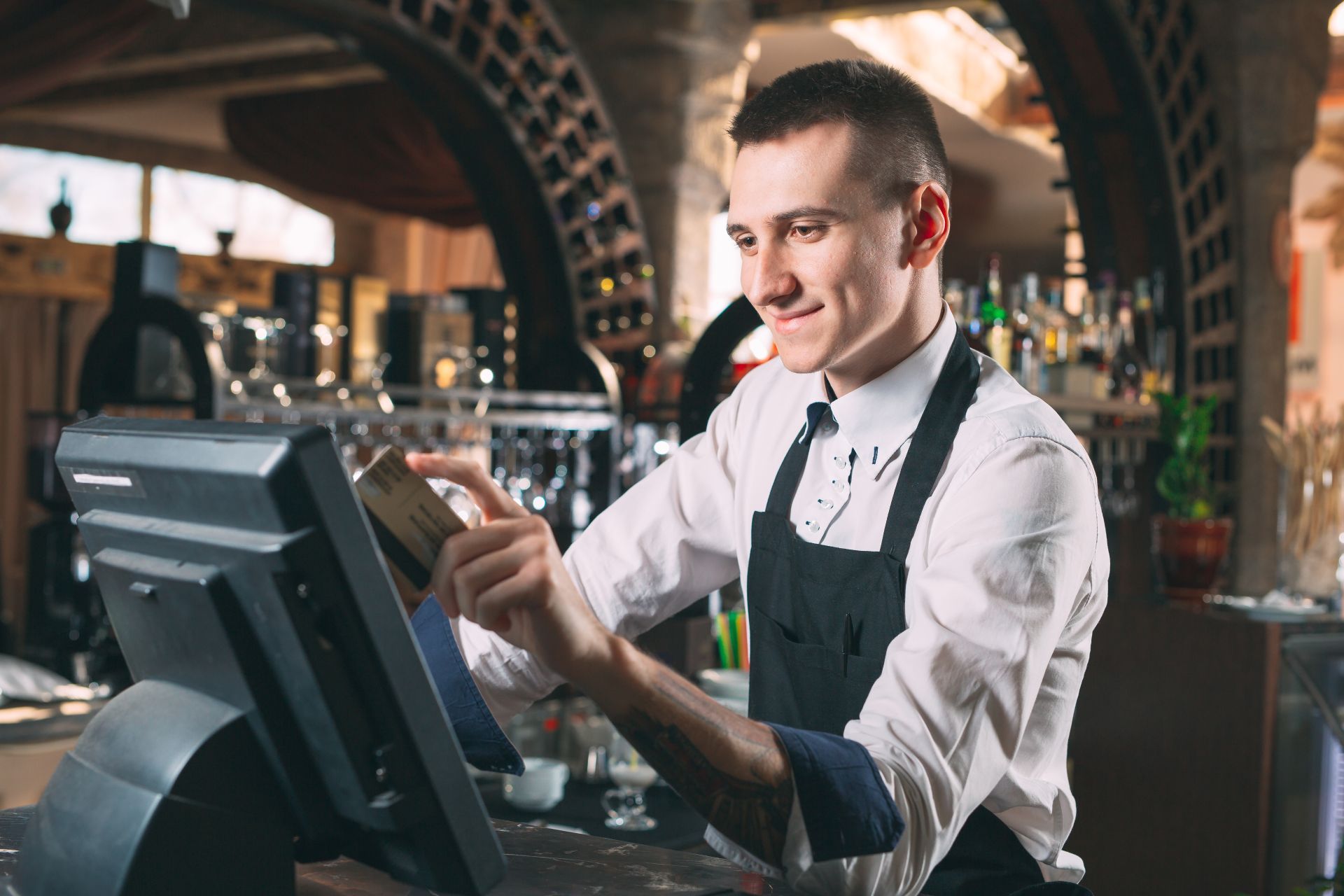 Bartender swipes a credit card at a POS system in a bar, wearing a white shirt, black apron, smiling.
