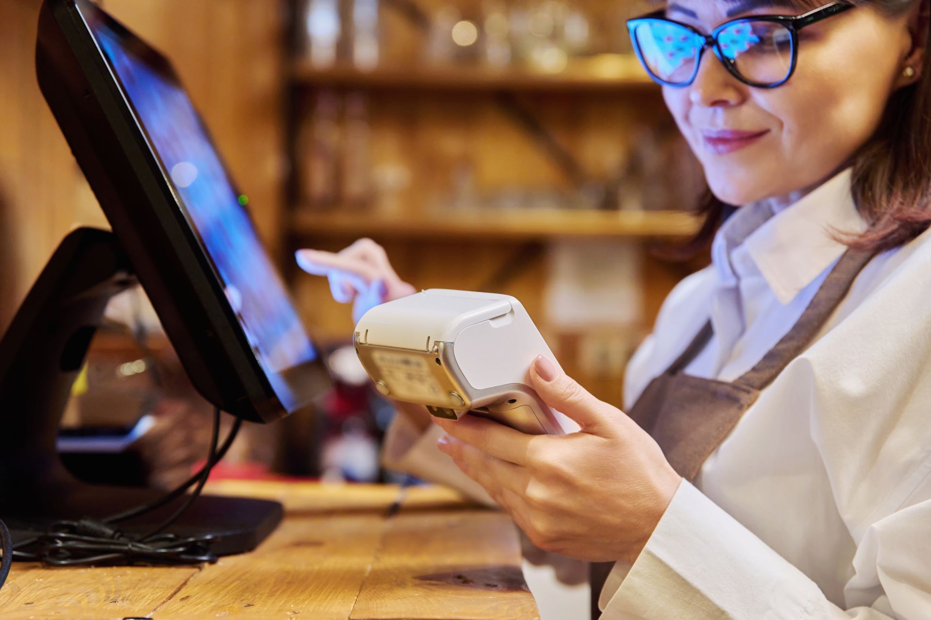 Woman in glasses using a card reader at a POS terminal, smiling. Wooden counter, neutral background.