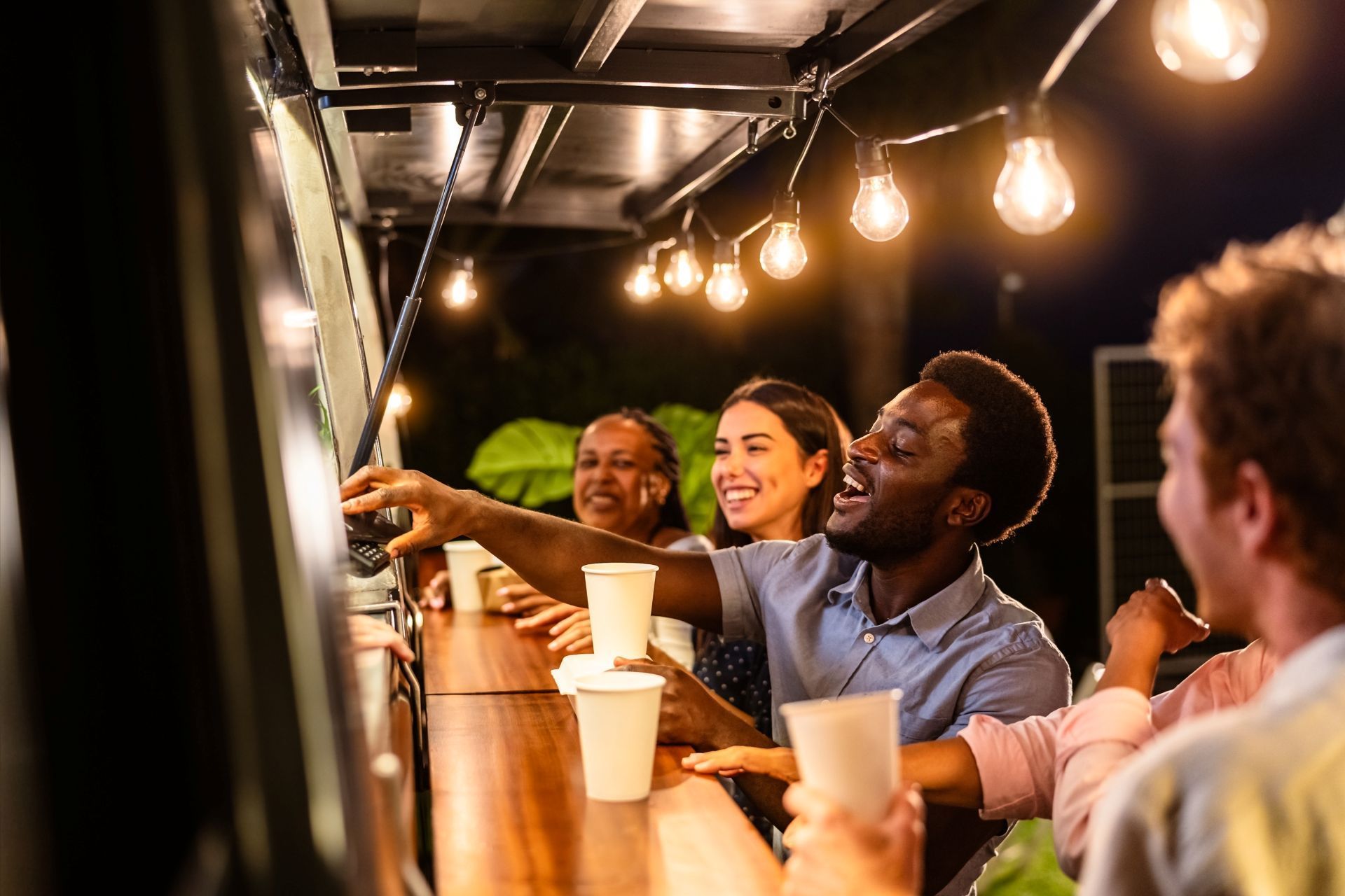 People at outdoor bar, lit by string lights, reaching for drinks.