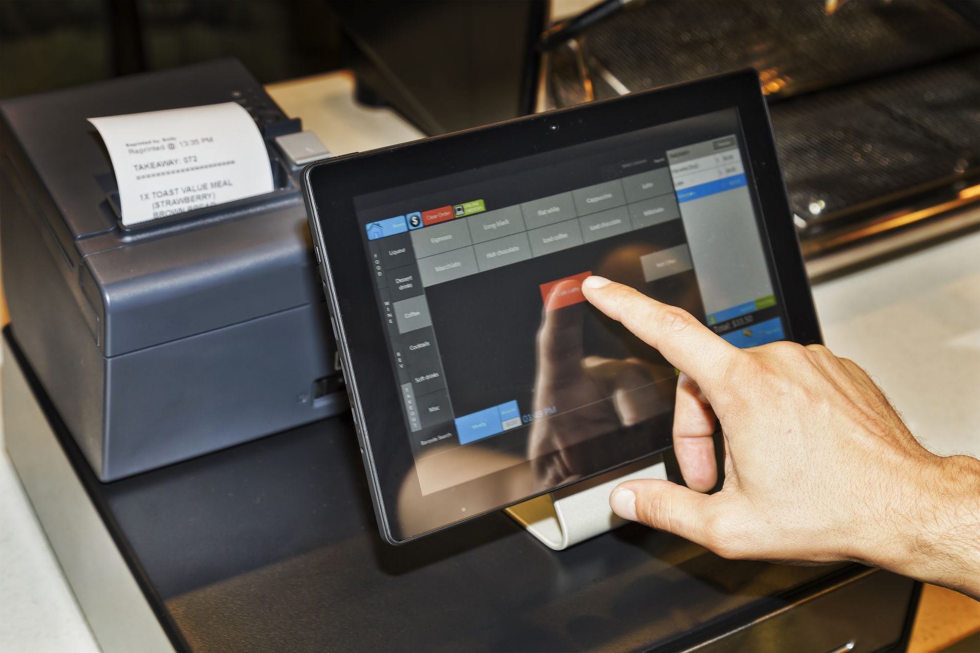Person's hand touching a tablet screen at a point-of-sale terminal. A receipt printer sits to the left.