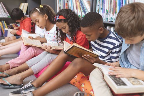 Children reading books on the floor in a library, surrounded by bookshelves.
