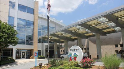 Children's hospital exterior with entry canopy, flag, and signage in front of a modern building on a sunny day.