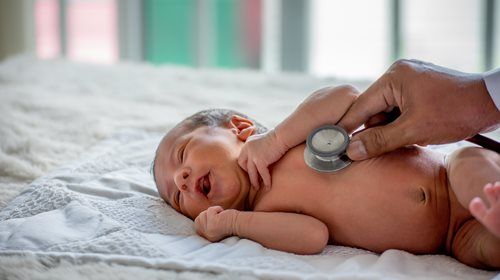 Doctor using a stethoscope to examine a newborn baby's chest. Baby is lying on a bed and smiling.