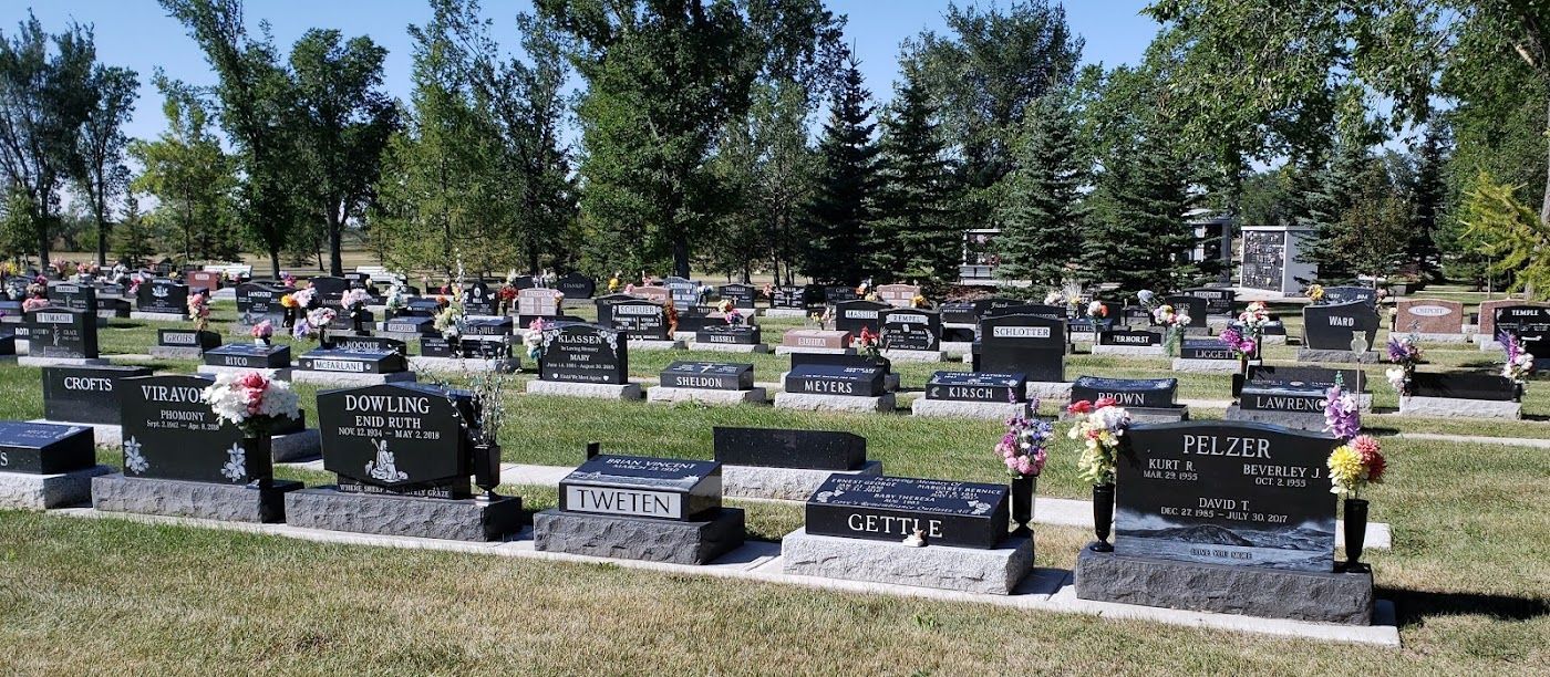 A cemetery with rows of headstones, trees in the background, under a bright blue sky.