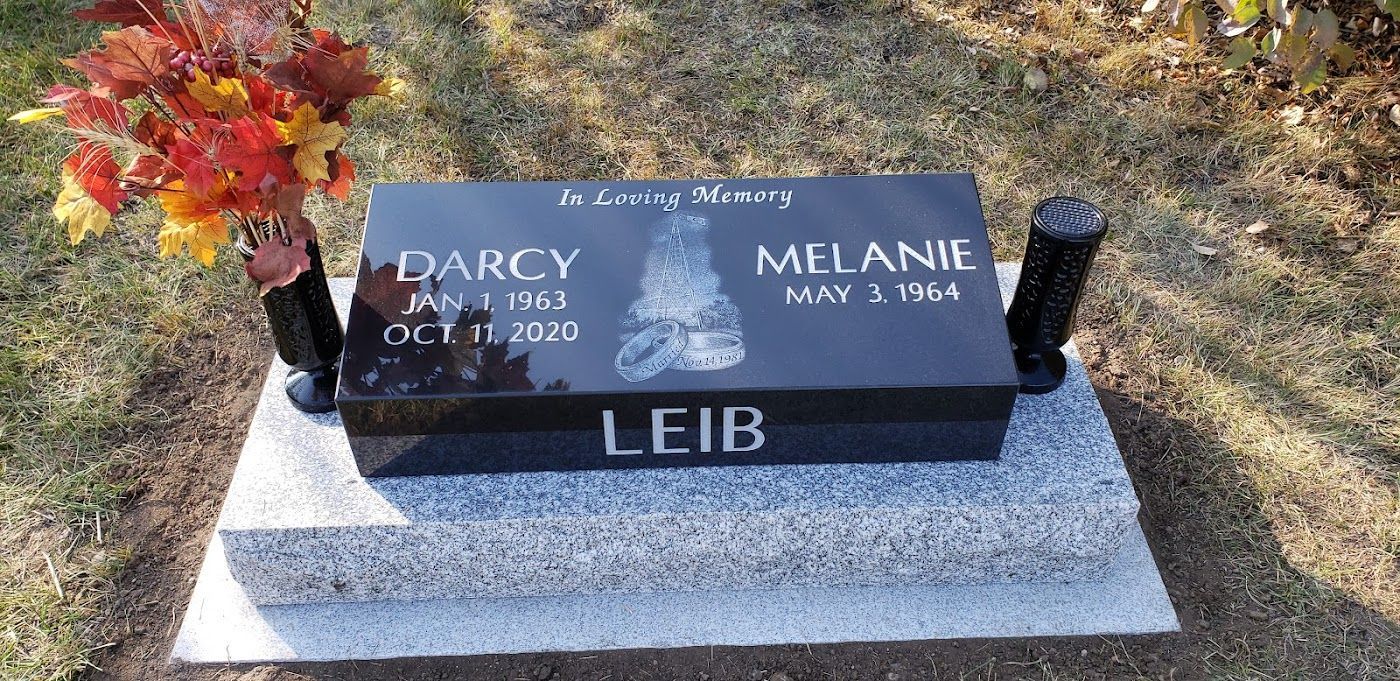Grave marker with two names: Darcy (Jan 1, 1963 - Oct 11, 2021) and Melanie (May 2, 1964), with flowers on it.