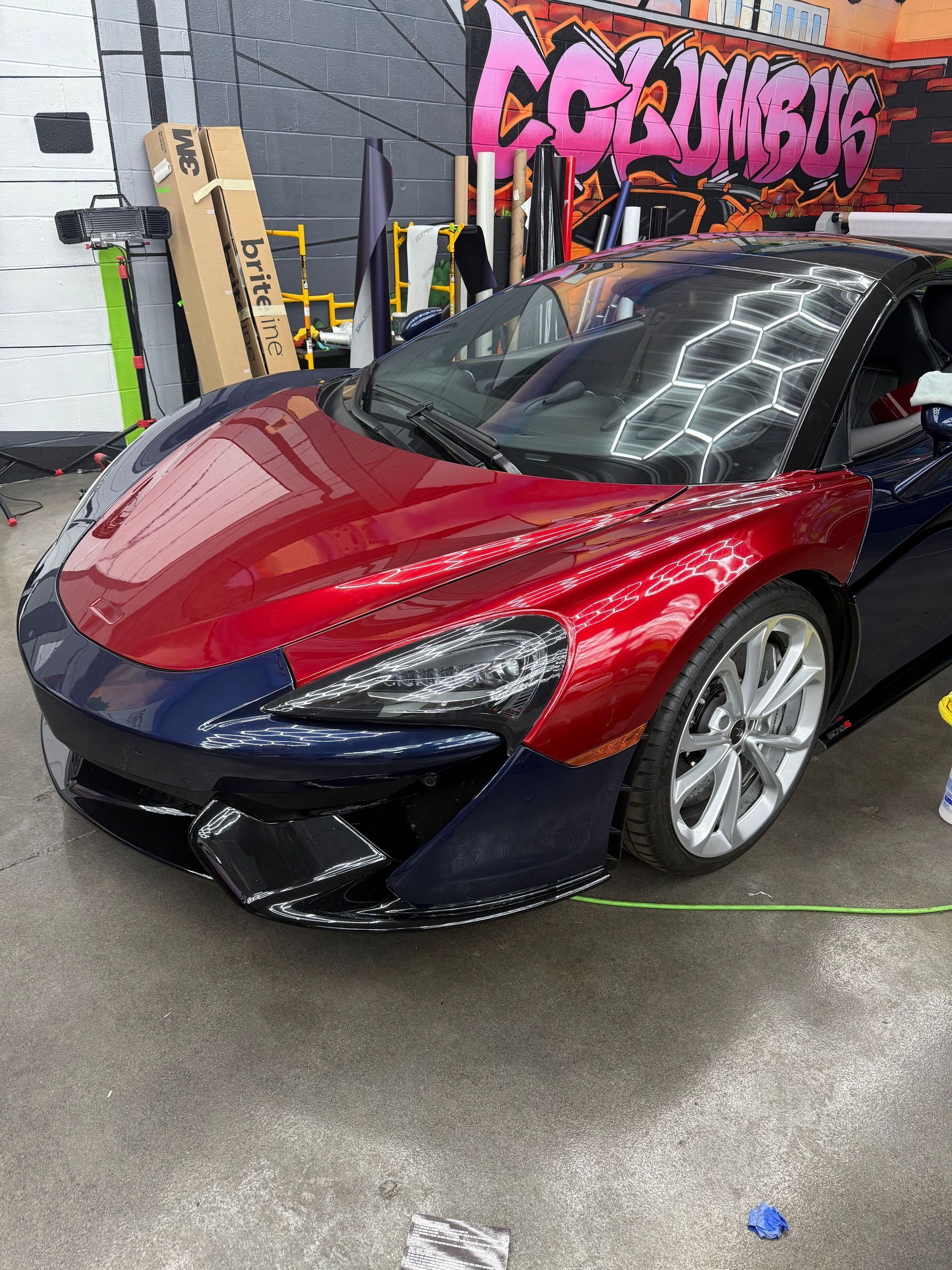 A blue and red McLaren sports car in a shop. The front is partially wrapped in red vinyl.
