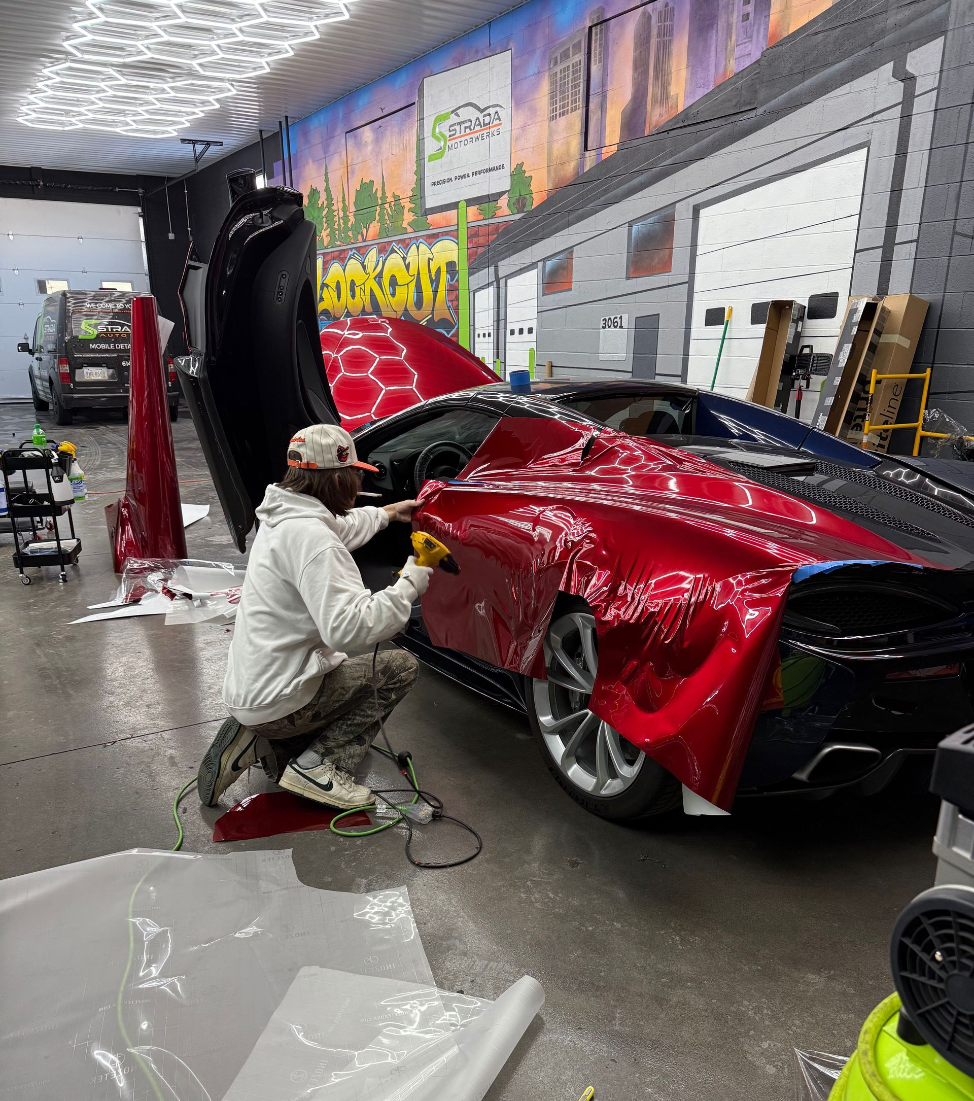 Man applying film to red sports car's side panel in a garage. Car door open, tools visible.