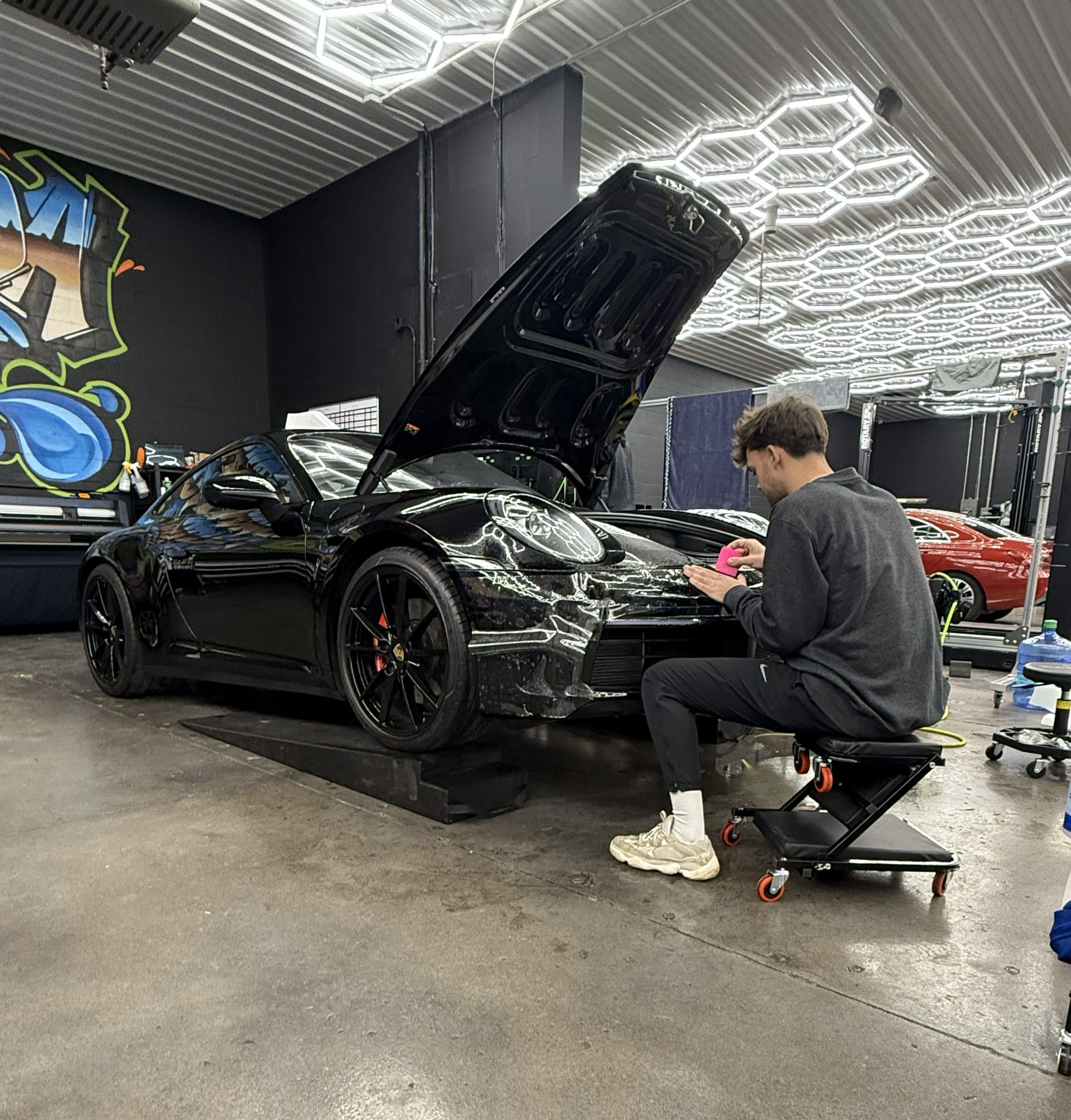 Man working on a black sports car with the hood open in a garage under bright lights.