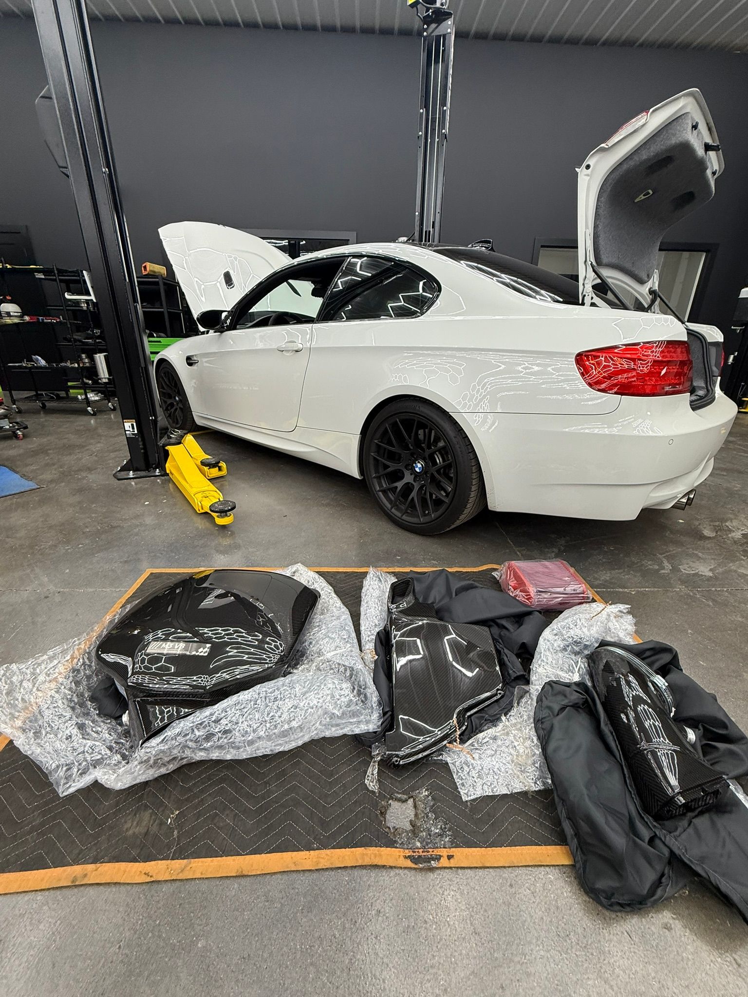 White car with open hood and trunk; carbon fiber parts laid out on the floor. Garage setting.