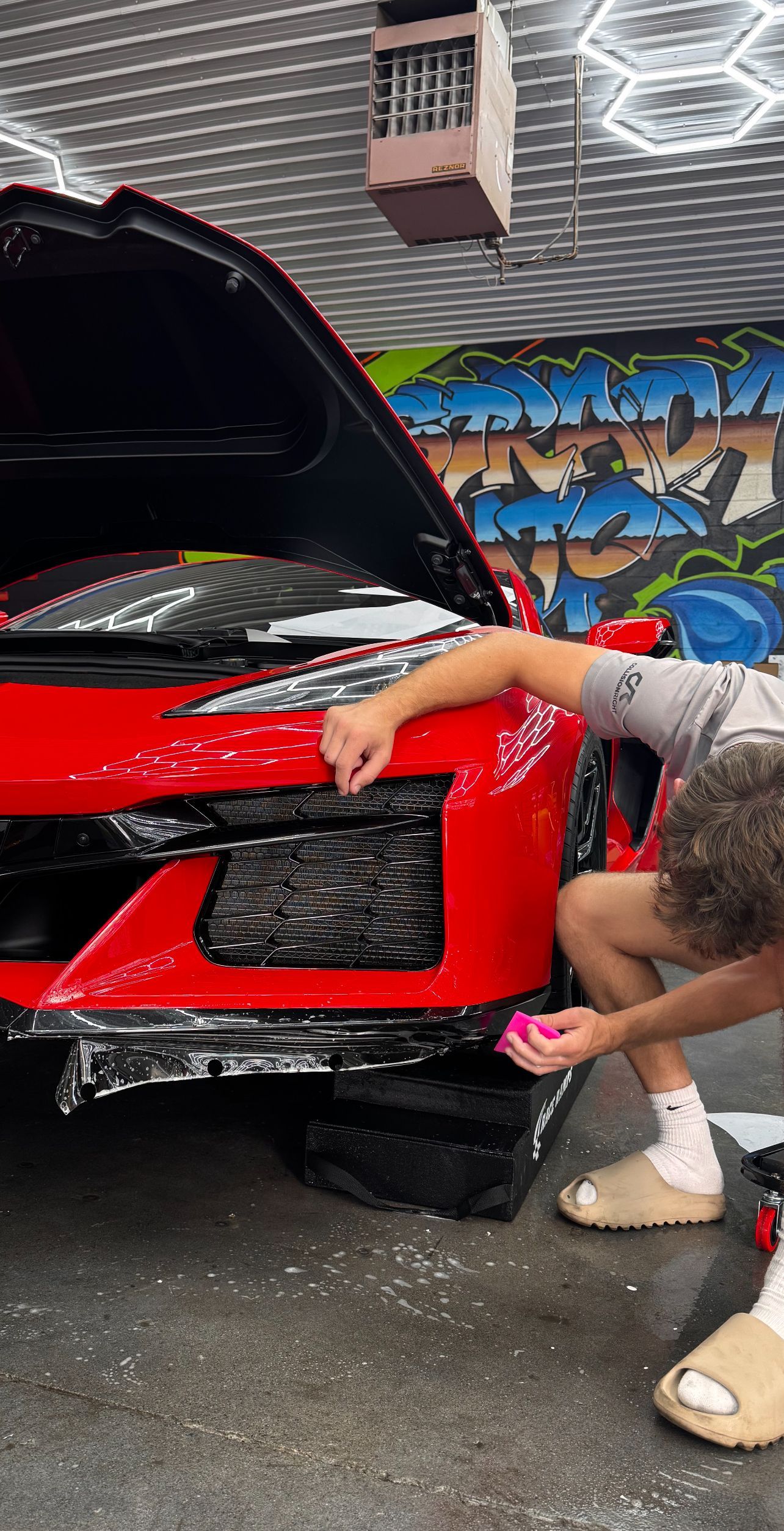A person in a gray shirt and tan slides applies a protective film to the front bumper of a red sports car in a garage.
