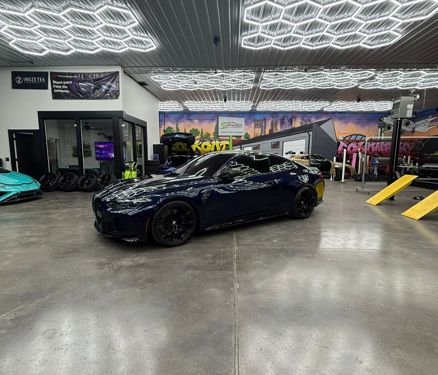 Blue car on a lift in a garage, with a cart of yellow suspension parts.