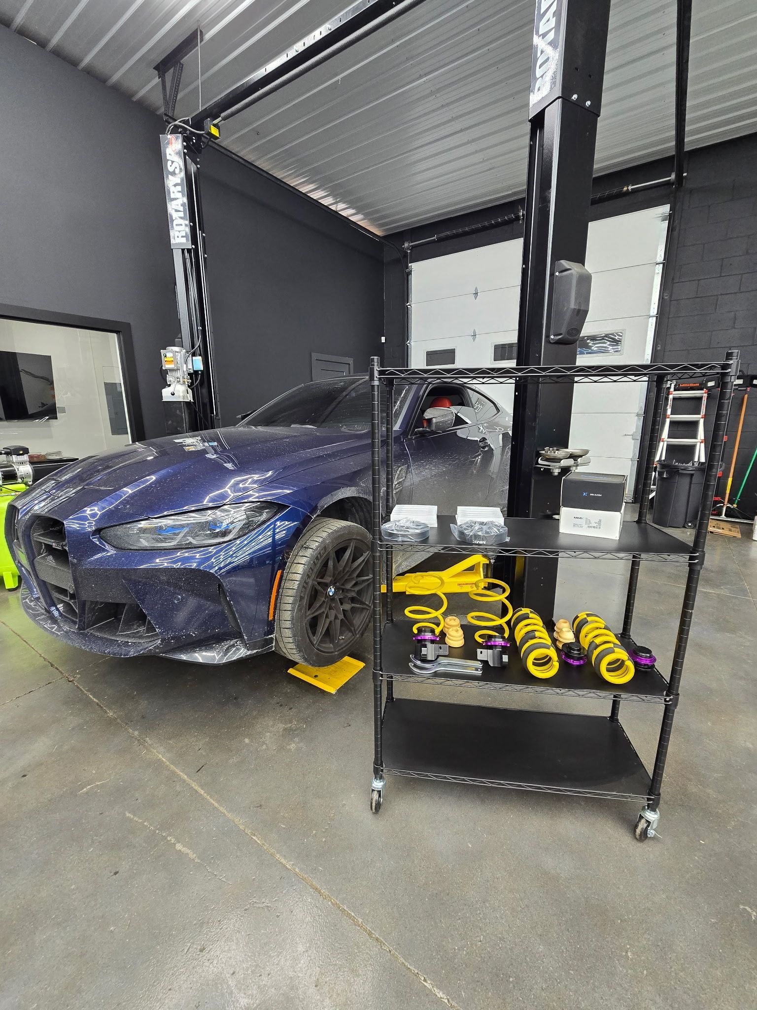 Blue car on a lift in a garage, with a cart of yellow suspension parts.