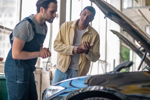 A mechanic in overalls and a customer in a yellow jacket discuss a repair with a car's hood open in a service garage.