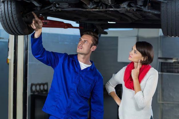 A mechanic in blue coveralls points to the underside of a raised car for a woman in a white sweater and red scarf.