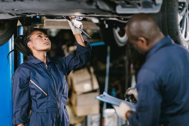 Two mechanics in blue uniforms work under a lifted car, with one using a wrench while the other holds a clipboard.