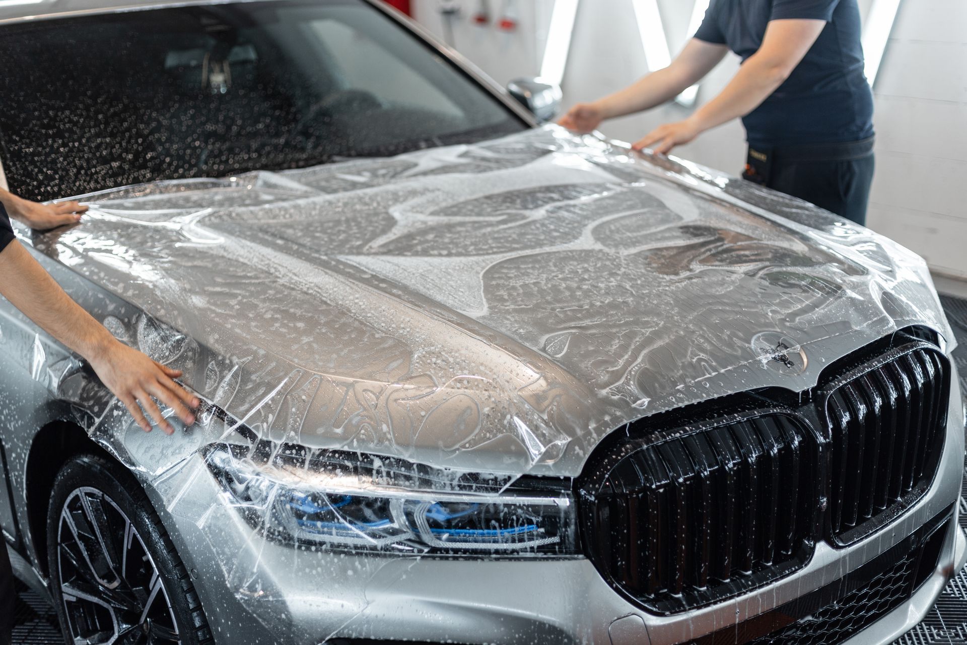 Two people apply a transparent protective film to the hood of a silver BMW in a workshop.