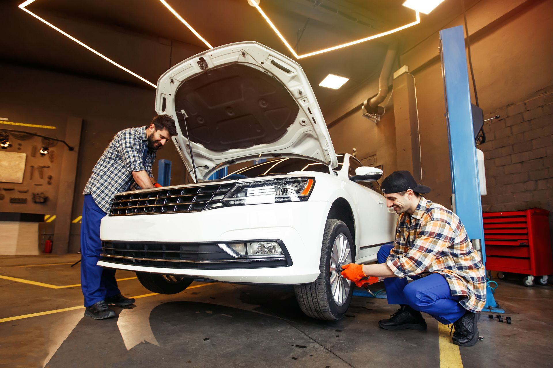 Two mechanics work on a white car in a garage; one checks the engine under an open hood while the other inspects a tire.
