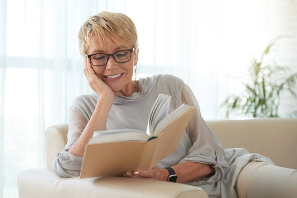 Woman wearing glasses smiles while reading a book on a beige couch near a window.