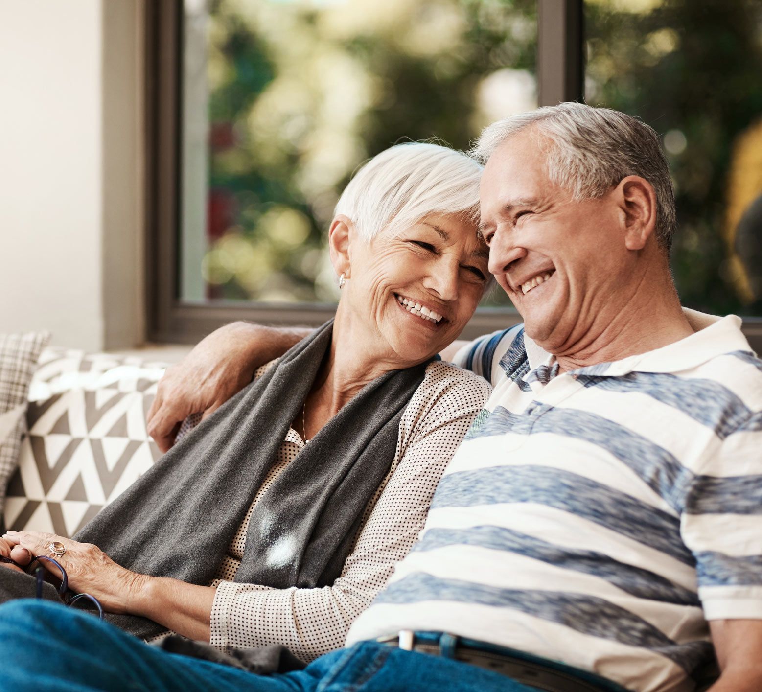 Smiling couple, arms around each other, sitting close together indoors, appearing happy.