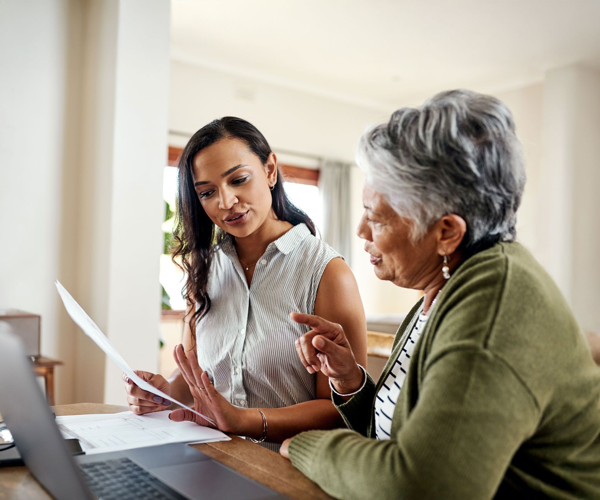 Woman showing documents to older person seated at a table with a laptop.