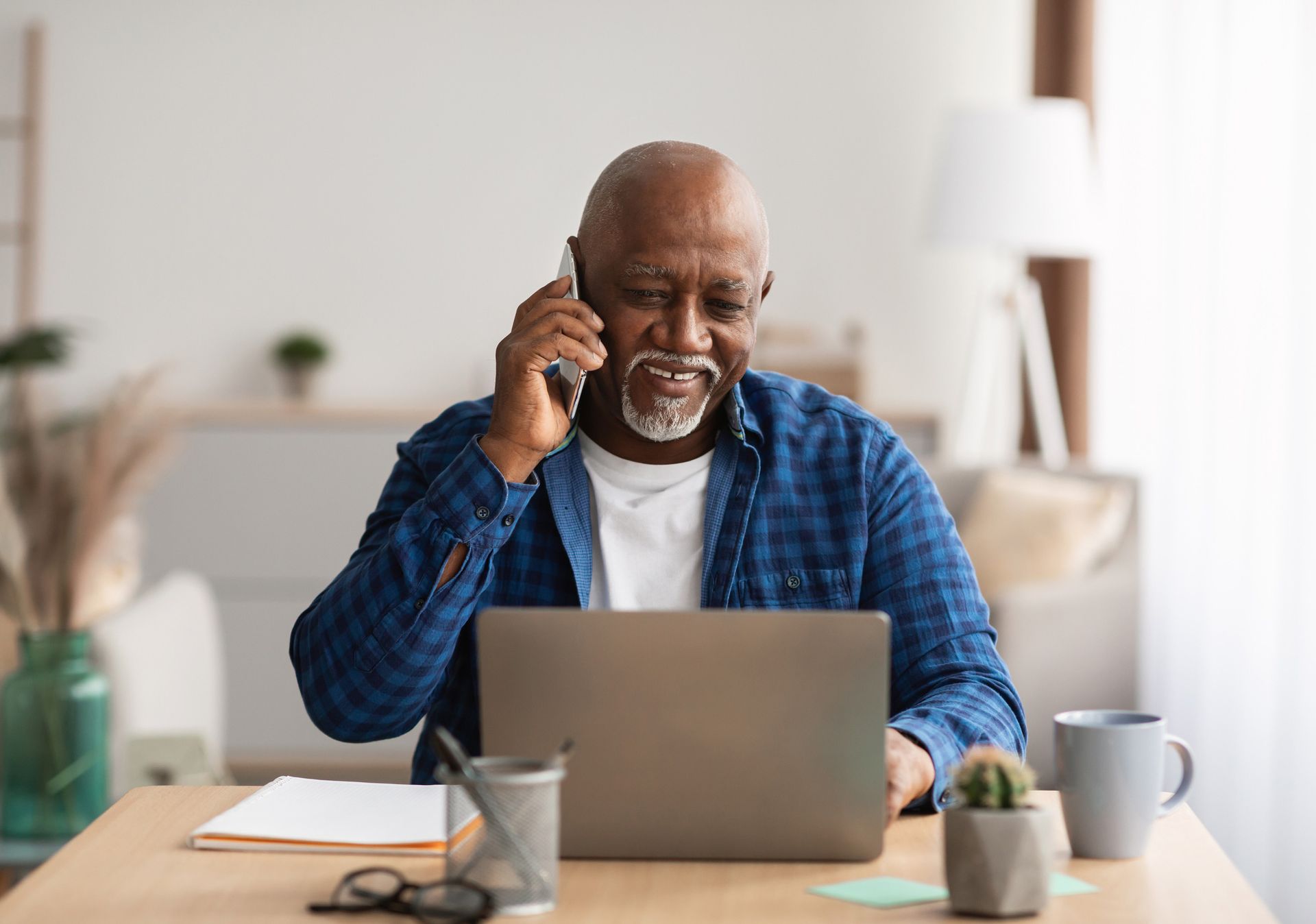 Man on the phone at a desk with a laptop, smiling. Neutral indoor setting with plants and a coffee cup.