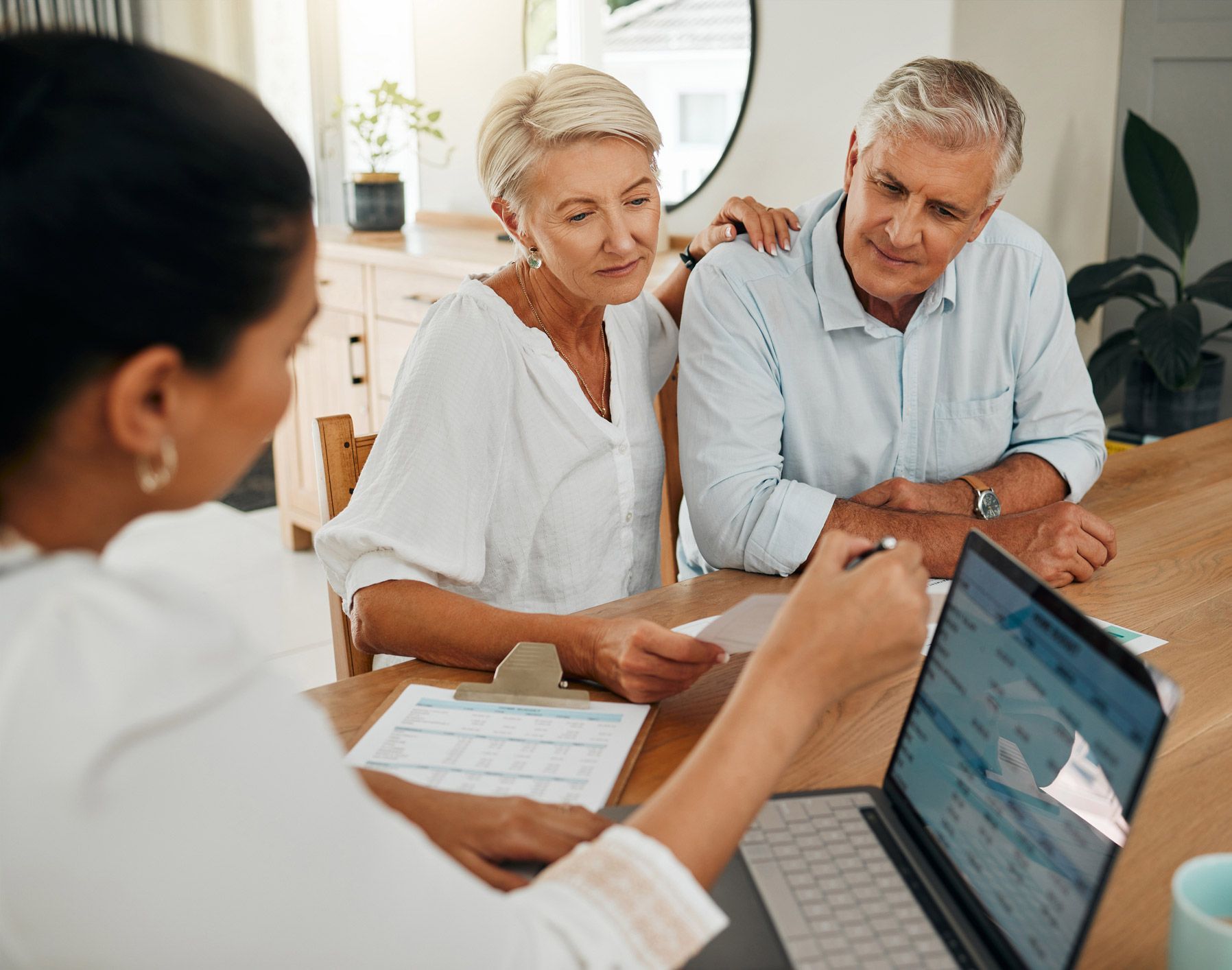 Woman advising a couple at a table, pointing at a laptop screen. They review documents, indoors.