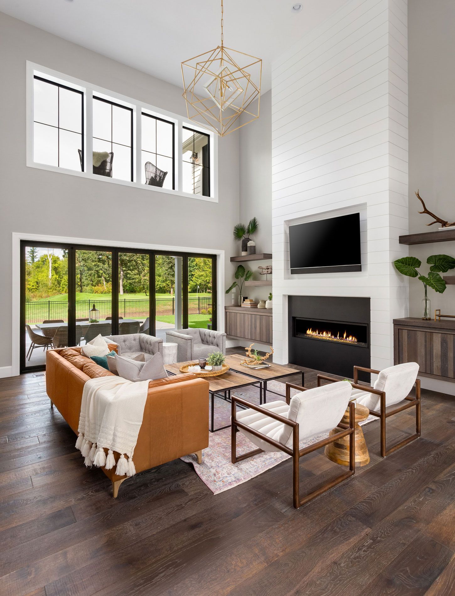 Modern living room with high ceilings, fireplace, and large windows overlooking a backyard.