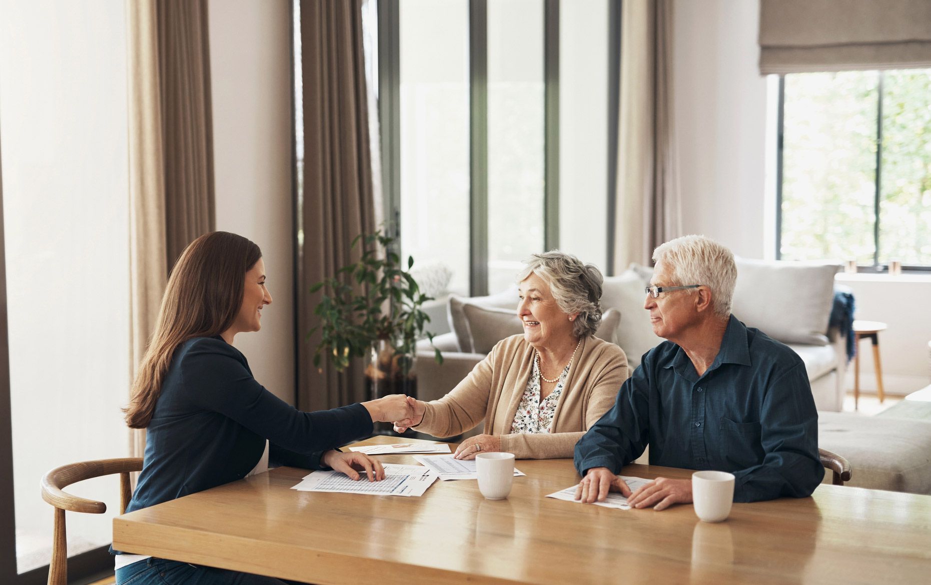 Woman shaking hands with a seated senior woman at a table with a senior man. Papers and coffee cups are present. Indoors.