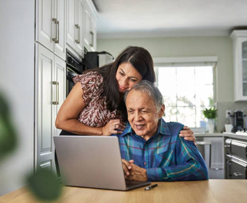 Woman assisting a senior man with a laptop in a kitchen, both smiling at the screen.