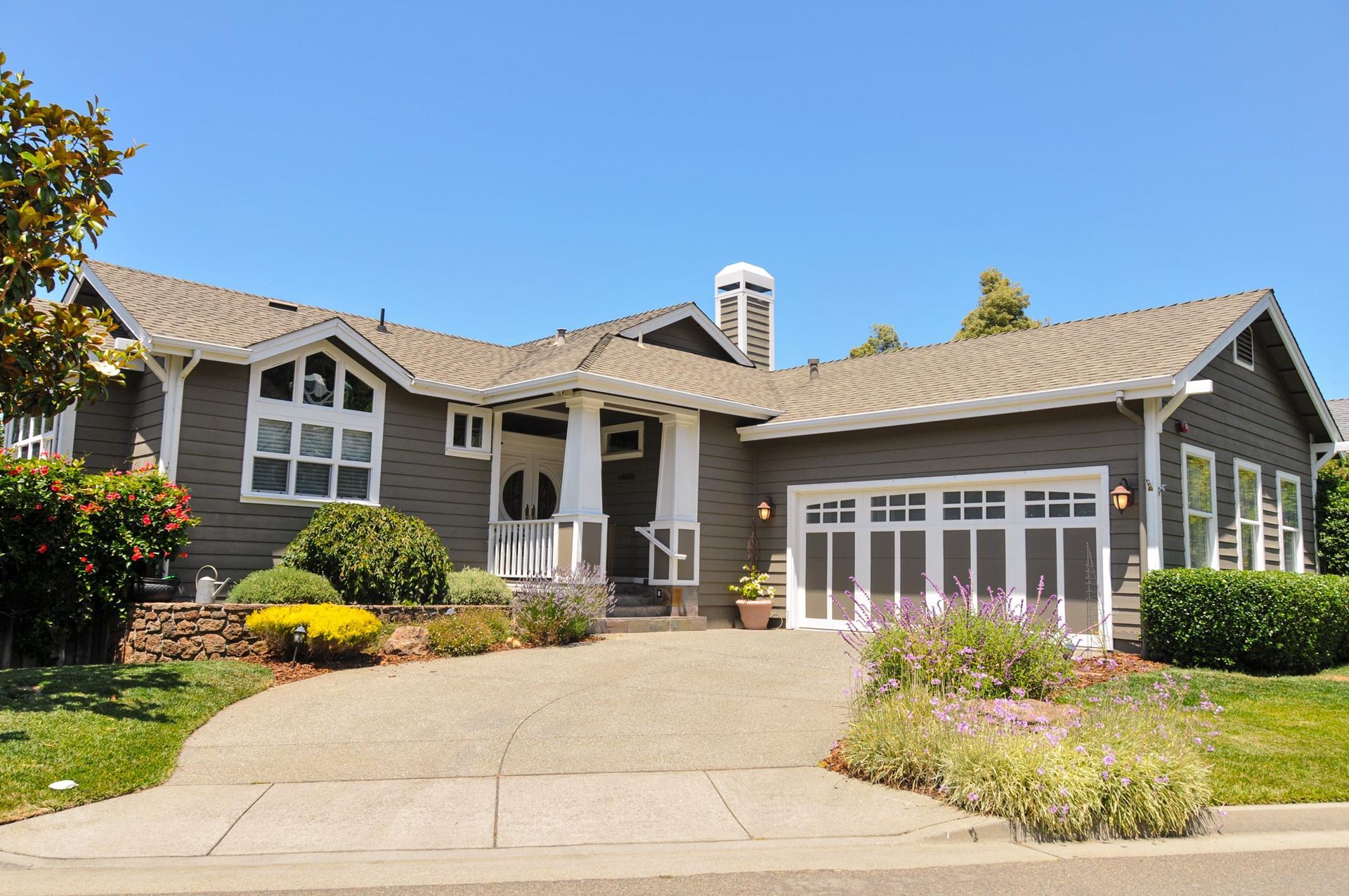 Gray house with white trim, arched windows, and a driveway. Landscaping and a blue sky are also visible.