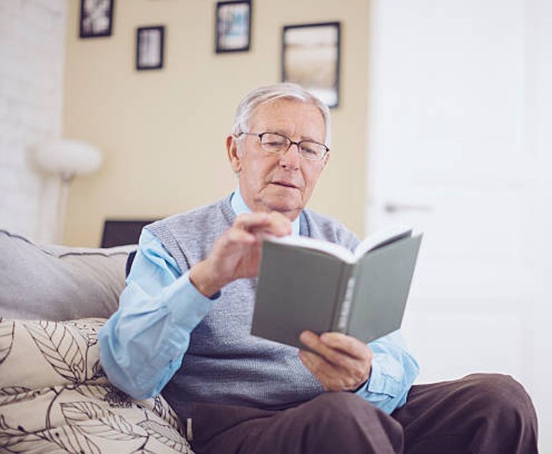 An elderly man in glasses reads a book on a couch in a living room, holding the book open.