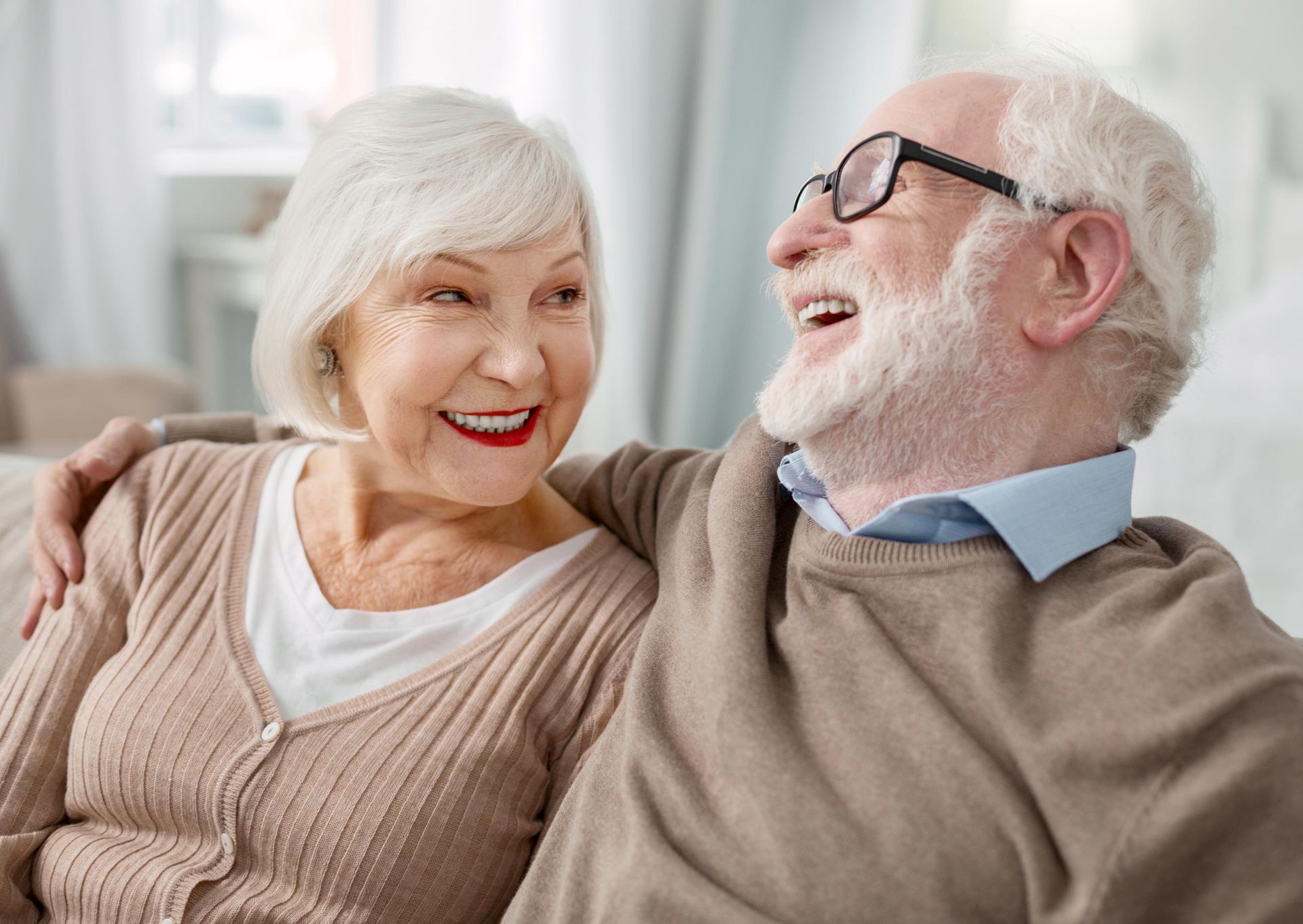 Elderly couple smiling and embracing indoors, man with glasses, woman with arm around him.