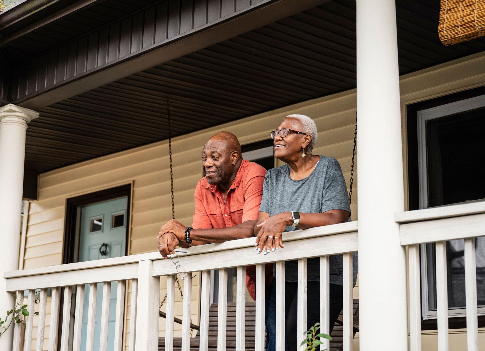 Couple on porch, smiling and looking off-camera. Leaning on white railing, beige house background.