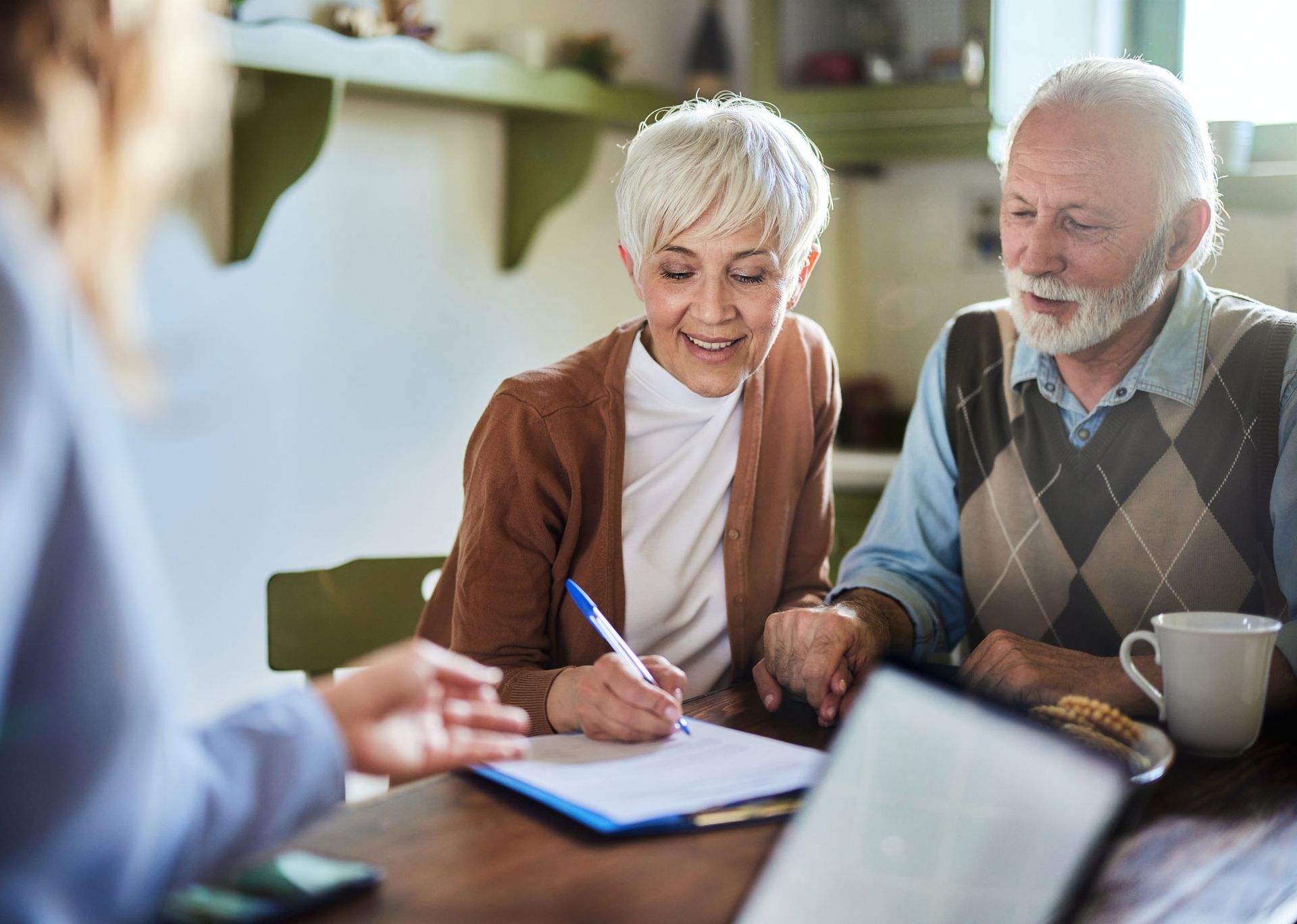 Senior couple signing paperwork with a person at a table, sunny interior.