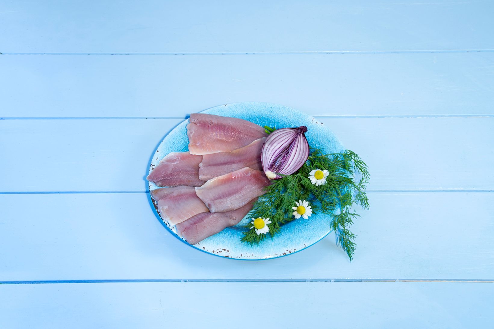 Ein blauer Teller mit Fischscheiben, Zwiebeln und Gänseblümchen auf einem Holztisch.