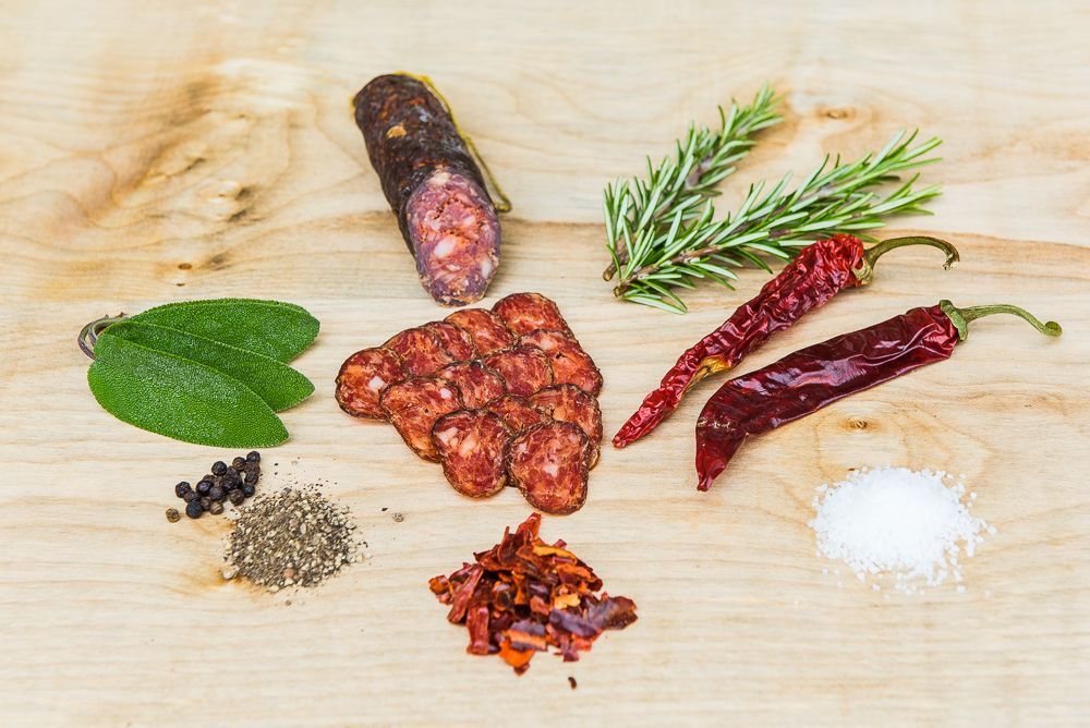 Ein hölzernes Schneidebrett mit Wurst, Paprika, Kräutern und Salz.