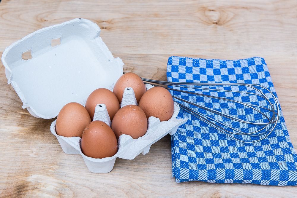 Auf einem Holztisch steht eine Schachtel Eier neben einem blau karierten Handtuch und einem Schneebesen.