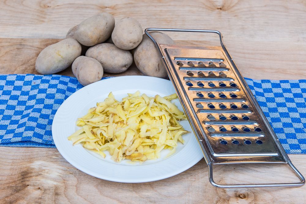 Ein Teller mit geriebenen Kartoffeln neben einer Reibe und Kartoffeln