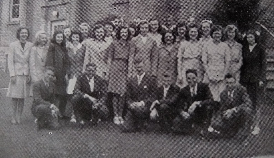 A black and white photo of a group of young people standing in front of a brick building.