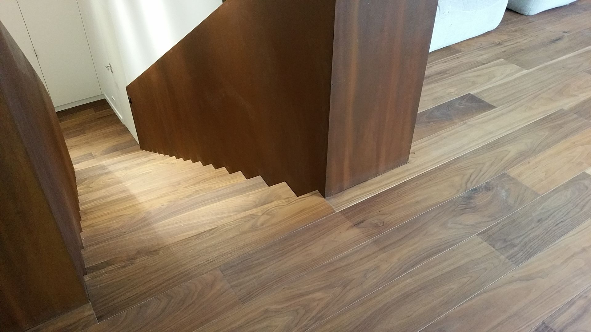 Escalier en bois descendant, avec un panneau latéral en bois foncé. Sol en bois brun.