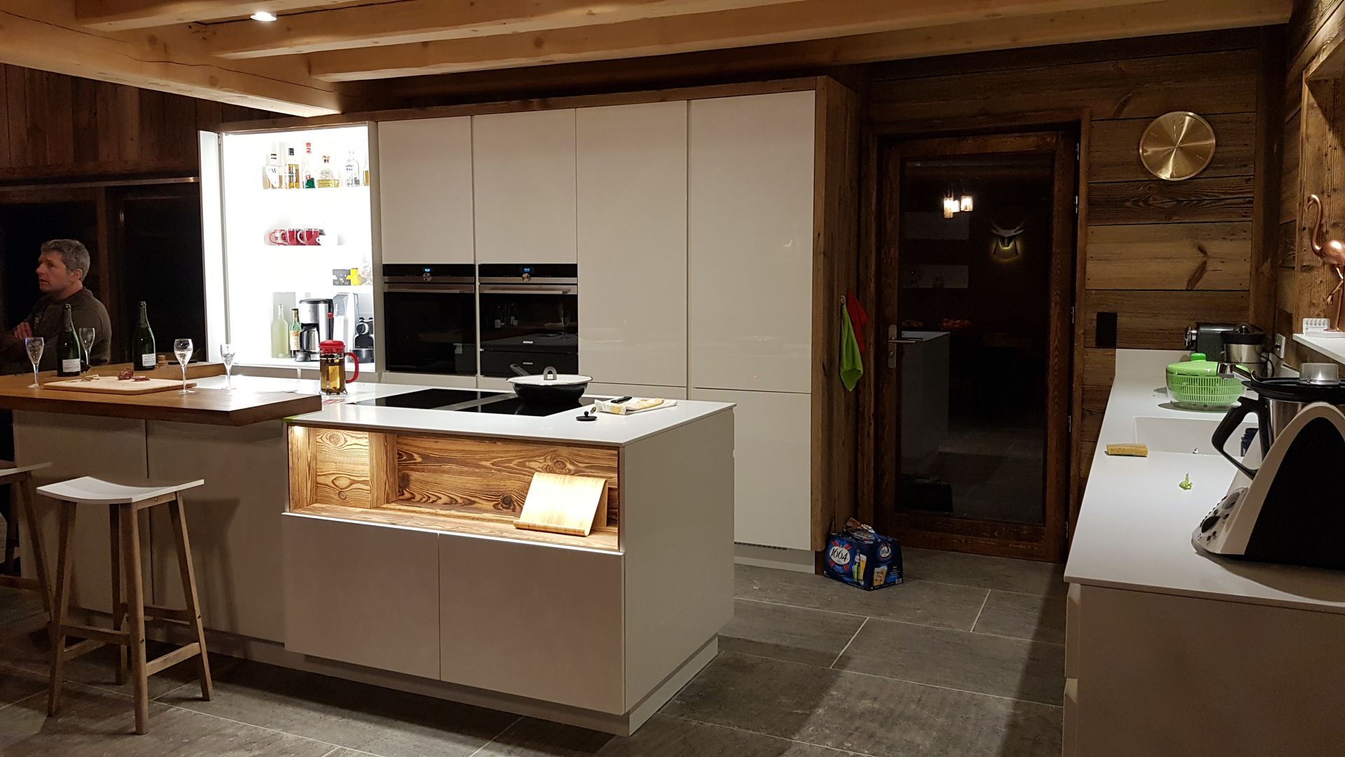 Cuisine moderne avec armoires blanches, îlot central et éléments en bois ; une personne se tient près de l'îlot.