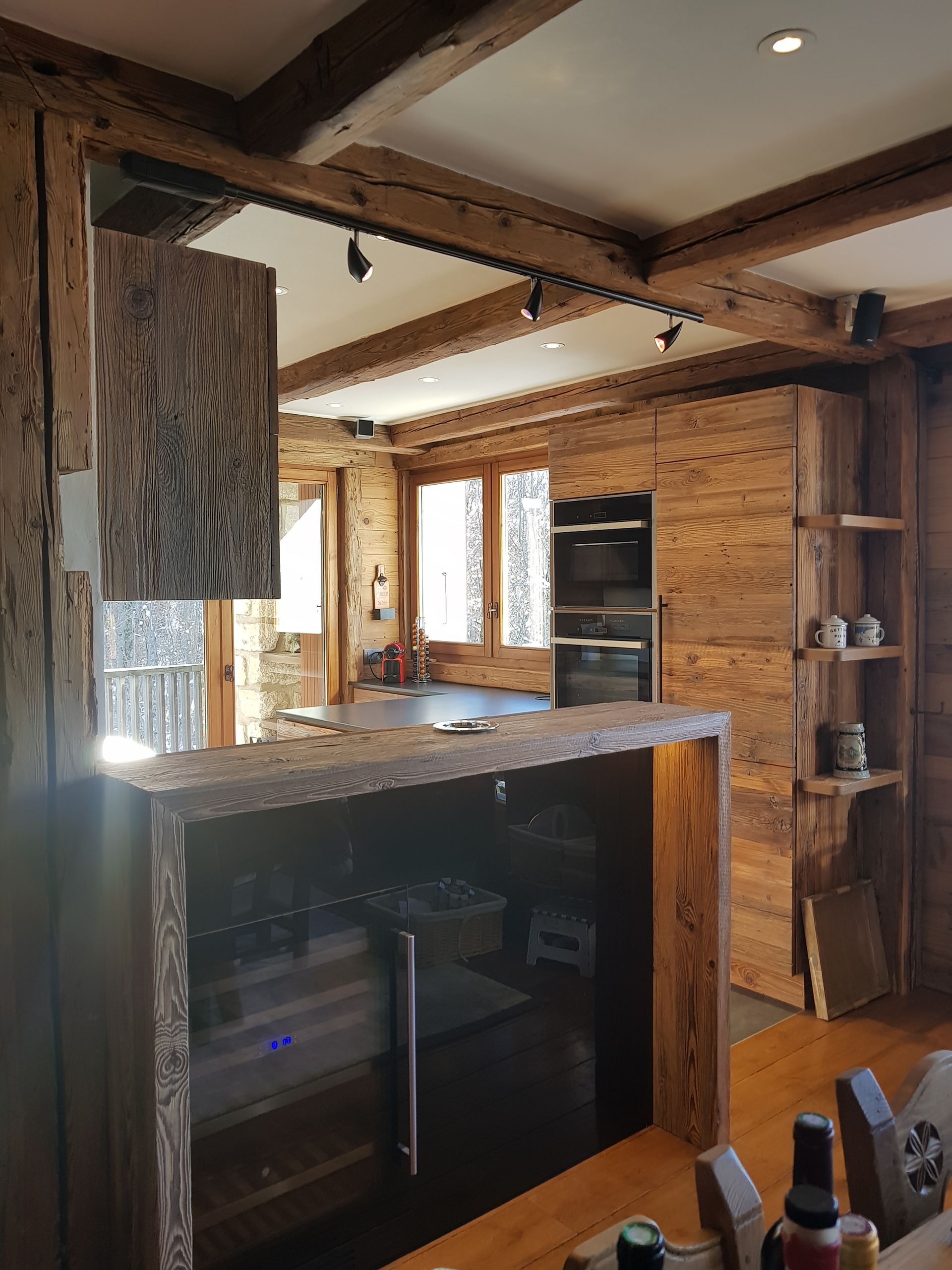 Cuisine en bois avec poutres apparentes, placards, bar et appareils électroménagers intégrés ; intérieur lumineux, journée ensoleillée.