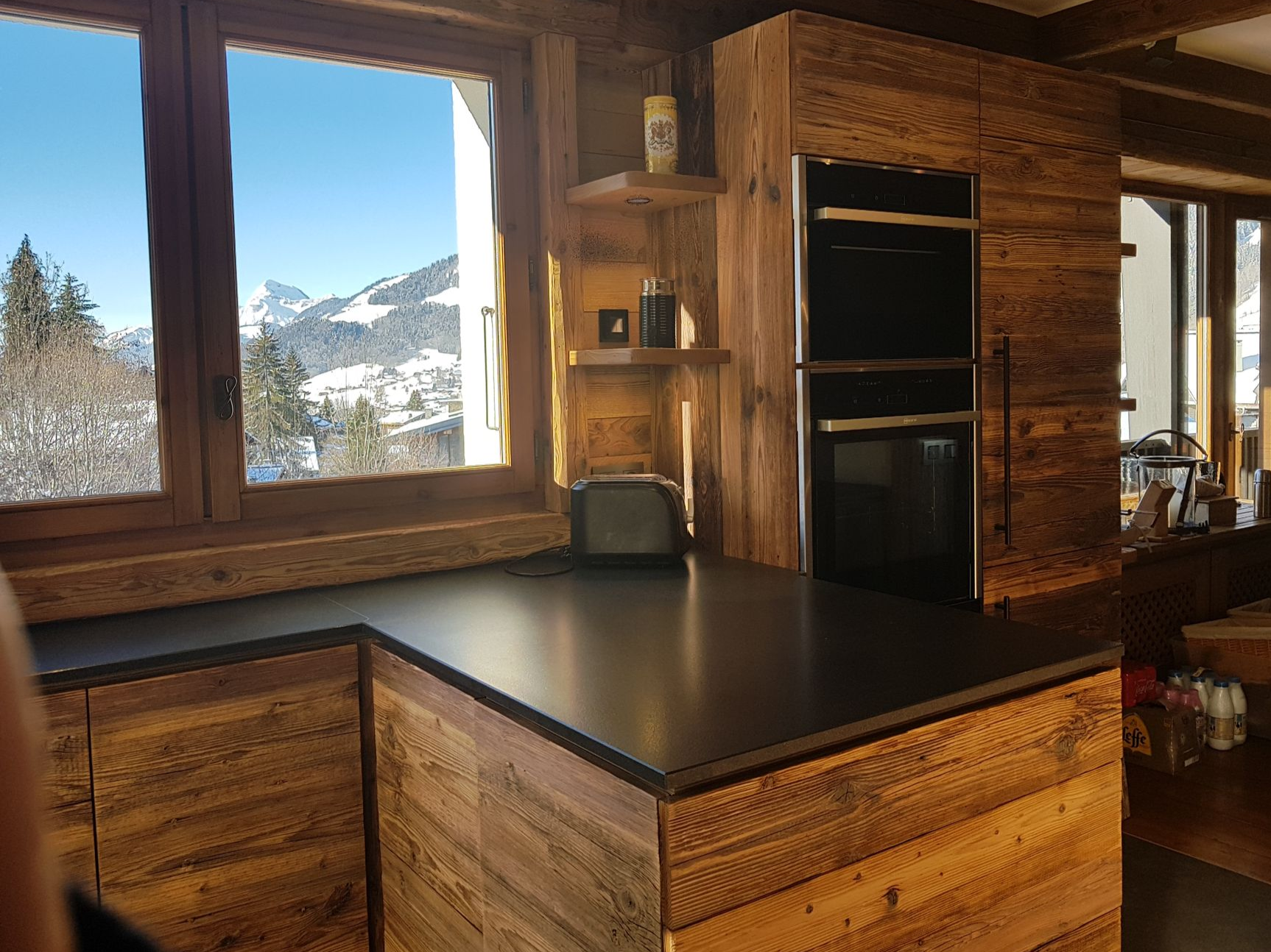 Cuisine avec placards en bois, plans de travail noirs et vue sur la montagne par la fenêtre.