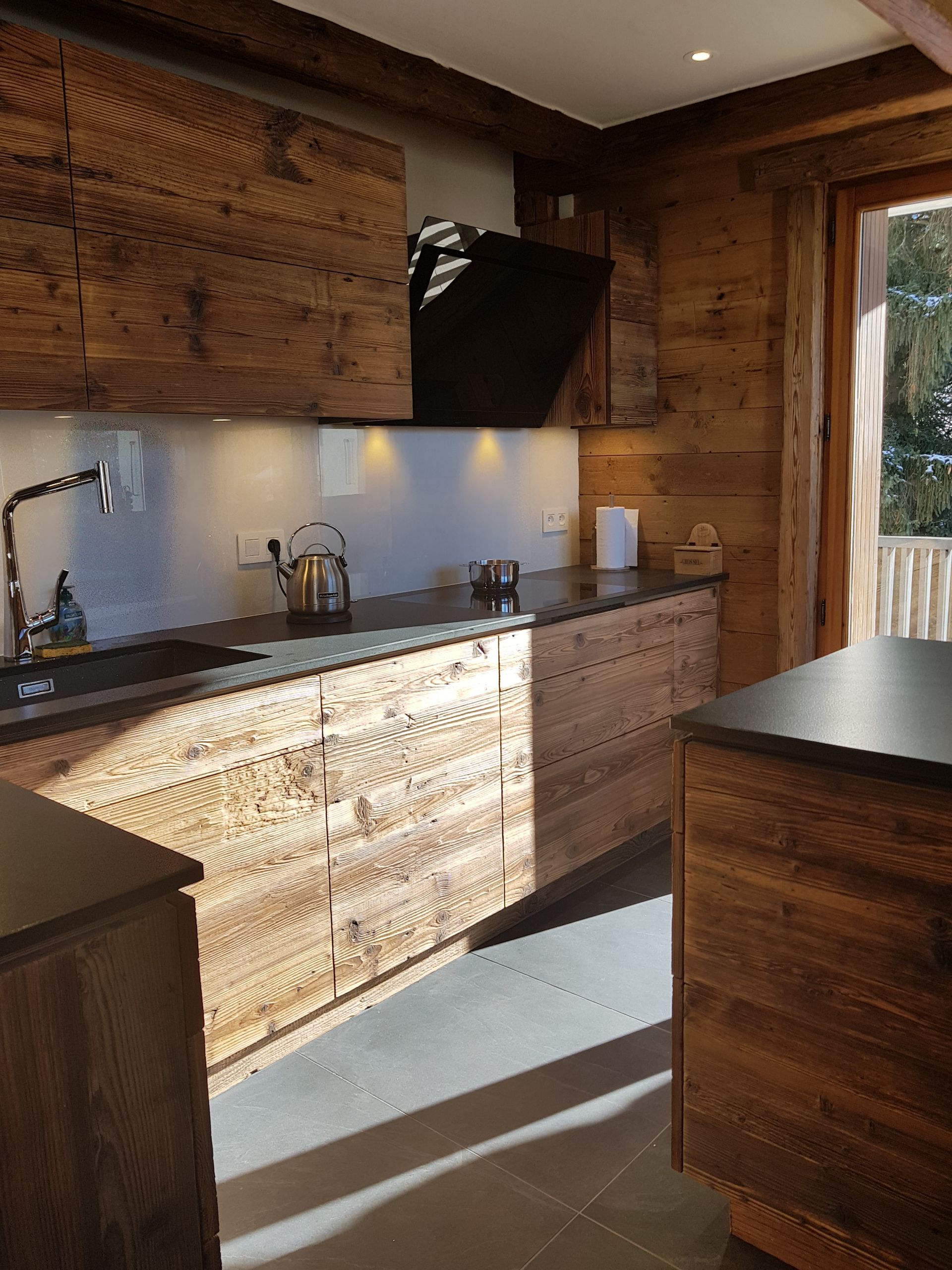 Cuisine en bois avec plans de travail, placards et murs foncés. Une fenêtre est visible en arrière-plan.